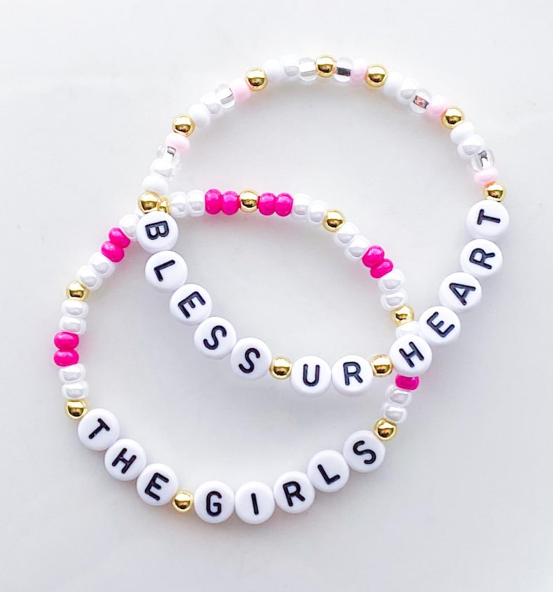 Puede incluir: Dos pulseras de cuentas con cuentas blancas, rosas y doradas. Una pulsera deletrea "BLESS UR HEART" y la otra dice "THE GIRLS" en letras negras sobre cuentas blancas. Un accesorio elegante.