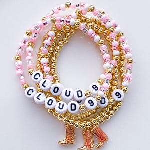 Puede incluir: Conjunto de pulseras de cuentas rosas, blancas y doradas. Las pulseras presentan el texto "CLOUD 9" en cuentas de letras blancas. Cuelgan dijes dorados de botas de vaquero.