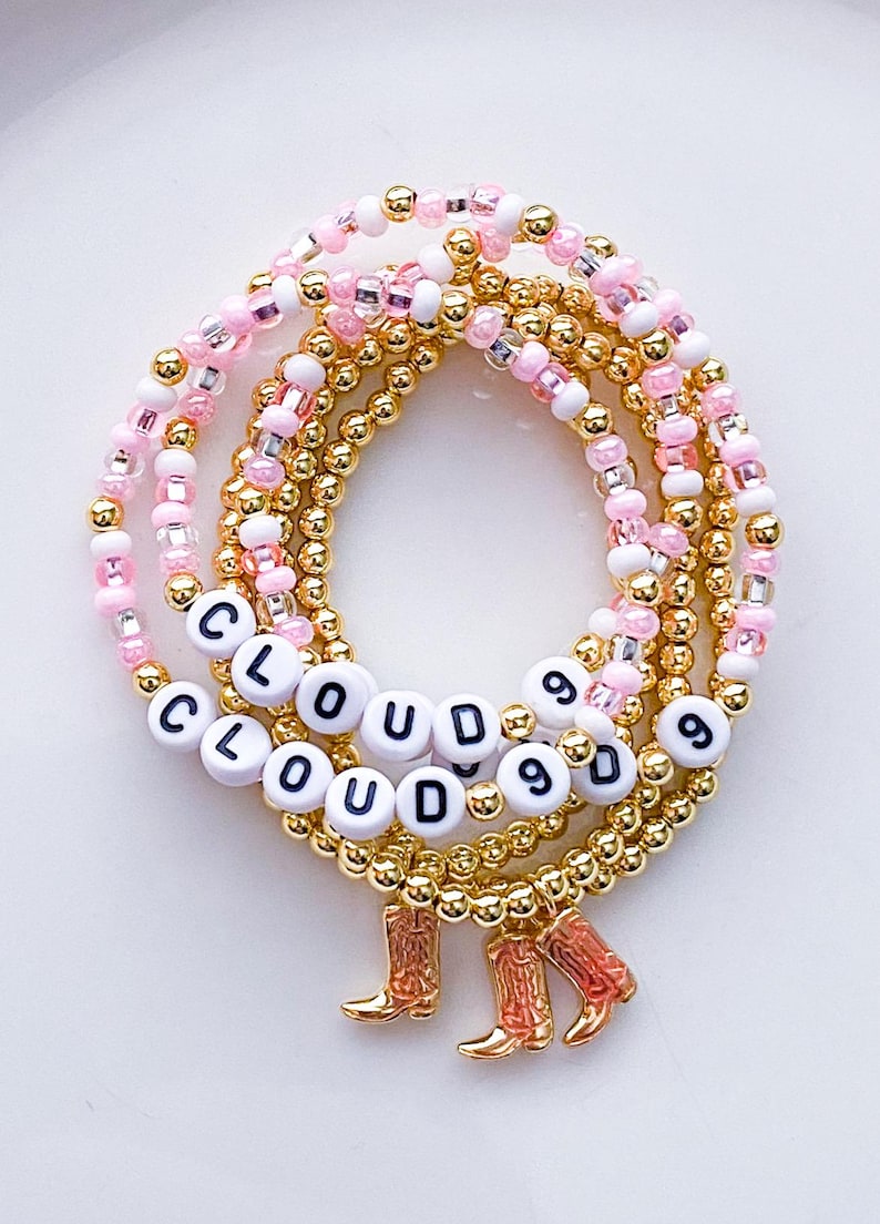 Puede incluir: Conjunto de pulseras de cuentas rosas, blancas y doradas. Las pulseras presentan el texto "CLOUD 9" en cuentas de letras blancas. Cuelgan dijes dorados de botas de vaquero.