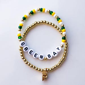 May include: Two beaded bracelets. One gold-colored bracelet with the word "GREEN BAY" spelled out in white letter beads. The other bracelet has green, yellow, and white beads. A gold-colored charm in the shape of a pizza slice hangs from the bottom bracelet.