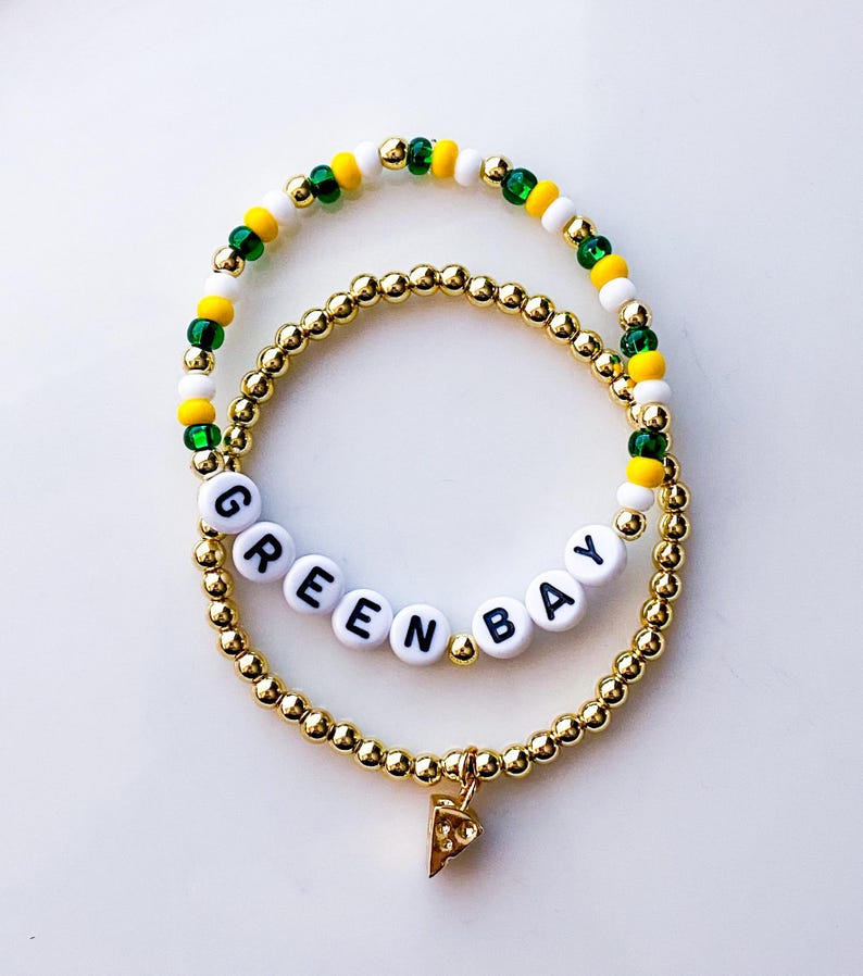 May include: Two beaded bracelets. One gold-colored bracelet with the word "GREEN BAY" spelled out in white letter beads. The other bracelet has green, yellow, and white beads. A gold-colored charm in the shape of a pizza slice hangs from the bottom bracelet.