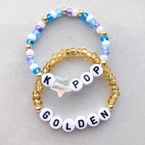 Pode incluir: Duas pulseiras de contas. Uma pulseira apresenta contas brancas com as letras "K POP" e "GOLDEN" em preto. Uma conta em forma de estrela est&aacute; entre as letras. A outra pulseira tem contas azuis, brancas e douradas.