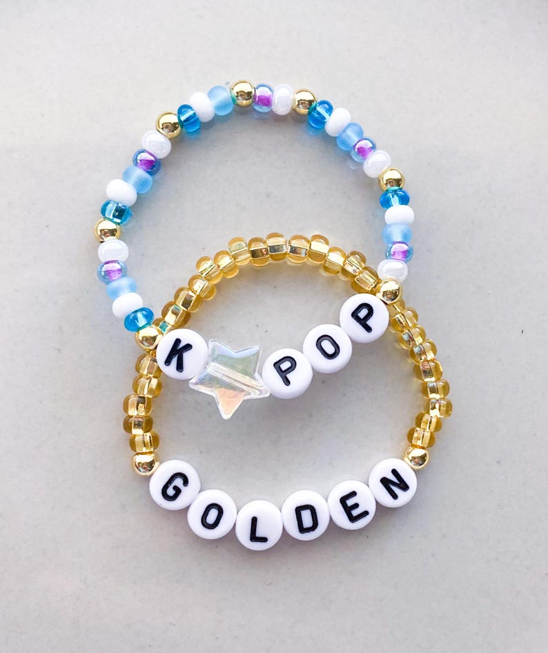 Pode incluir: Duas pulseiras de contas. Uma pulseira apresenta contas brancas com as letras "K POP" e "GOLDEN" em preto. Uma conta em forma de estrela est&aacute; entre as letras. A outra pulseira tem contas azuis, brancas e douradas.