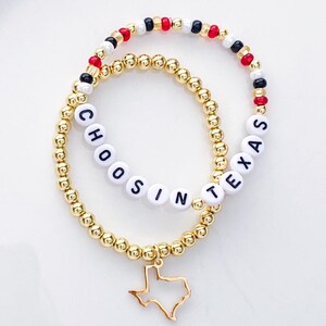 Puede incluir: Dos pulseras de cuentas doradas. Una pulsera deletrea "CHOOSIN TEXAS" con cuentas de letras blancas. La otra tiene cuentas rojas, negras y blancas. Un dije dorado en forma de Texas cuelga de la pulsera "CHOOSIN TEXAS".