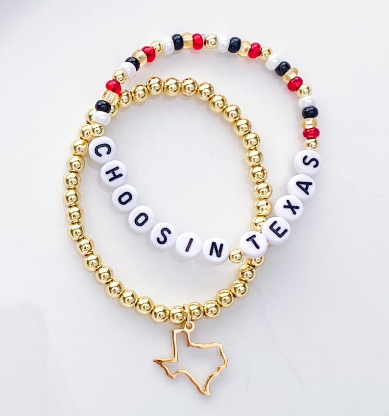 Puede incluir: Dos pulseras de cuentas doradas. Una pulsera deletrea "CHOOSIN TEXAS" con cuentas de letras blancas. La otra tiene cuentas rojas, negras y blancas. Un dije dorado en forma de Texas cuelga de la pulsera "CHOOSIN TEXAS".