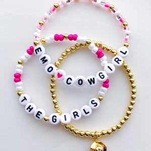 Puede incluir: Tres pulseras de cuentas. Una pulsera tiene el texto "EMO GIRL", otra "COWGIRL" con un coraz&oacute;n, y la tercera dice "THE GIRLS" con un dije dorado. Las cuentas son doradas, blancas, rosas y transparentes.