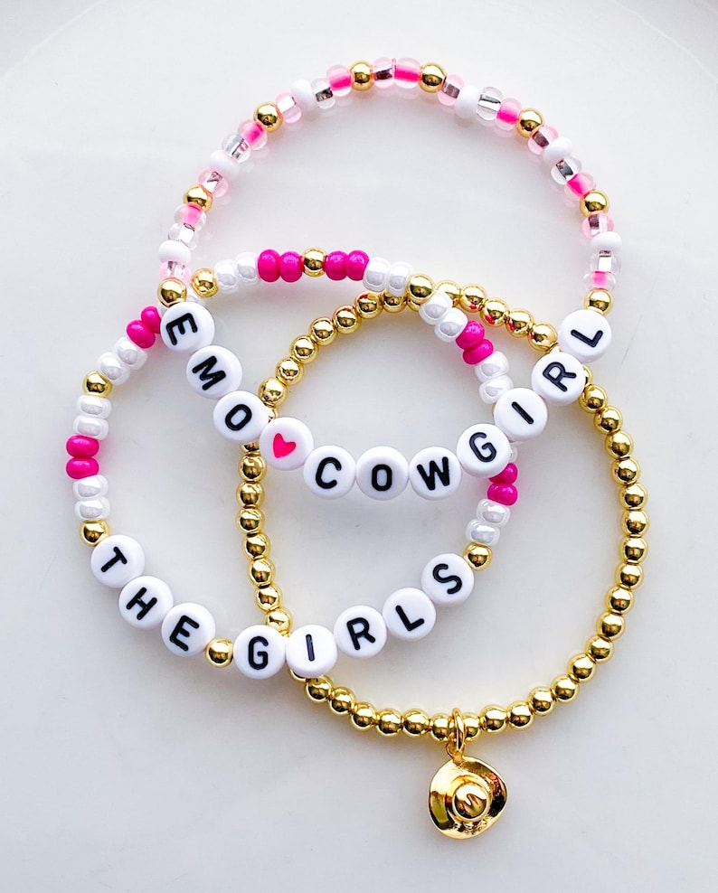 Puede incluir: Tres pulseras de cuentas. Una pulsera tiene el texto "EMO GIRL", otra "COWGIRL" con un coraz&oacute;n, y la tercera dice "THE GIRLS" con un dije dorado. Las cuentas son doradas, blancas, rosas y transparentes.