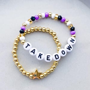 Pode incluir: Duas pulseiras de contas. Uma &eacute; dourada com a palavra "TAKE DOWN" escrita em contas de letras brancas. A outra pulseira &eacute; dourada com contas pretas, roxas e brancas. Um berloque de estrela dourada est&aacute; na pulseira "TAKE DOWN".