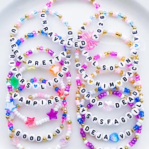 May include: A collection of beaded bracelets featuring white letter beads spelling phrases like UNRAVELED, PRETTY, DRIVER'S LICENSE, VAMPIRE, GOOD 4 U, SOUR, 2026, GIRLS SO IN, HIM BACK, DROP DEAD, SPSFAGSIL, DEJA VU, and GUTS, accented with colorful star and heart beads.