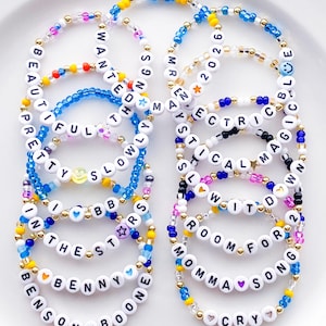 May include: A collection of beaded bracelets with white letter beads spelling out words and phrases like "BEAUTIFUL," "WANTED," and "ROOM FOR." The bracelets feature colorful beads in blue, yellow, pink, and gold, creating a vibrant and personalized accessory.