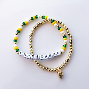 May include: Two beaded bracelets. One bracelet features the word "CHEESEHEAD" spelled out in white letter beads. The other bracelet is gold with a charm shaped like a cheese wedge. The bracelets have green, yellow, and white beads.