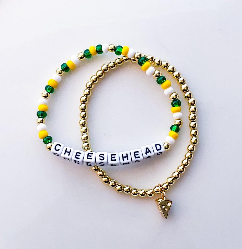 May include: Two beaded bracelets. One bracelet features the word "CHEESEHEAD" spelled out in white letter beads. The other bracelet is gold with a charm shaped like a cheese wedge. The bracelets have green, yellow, and white beads.
