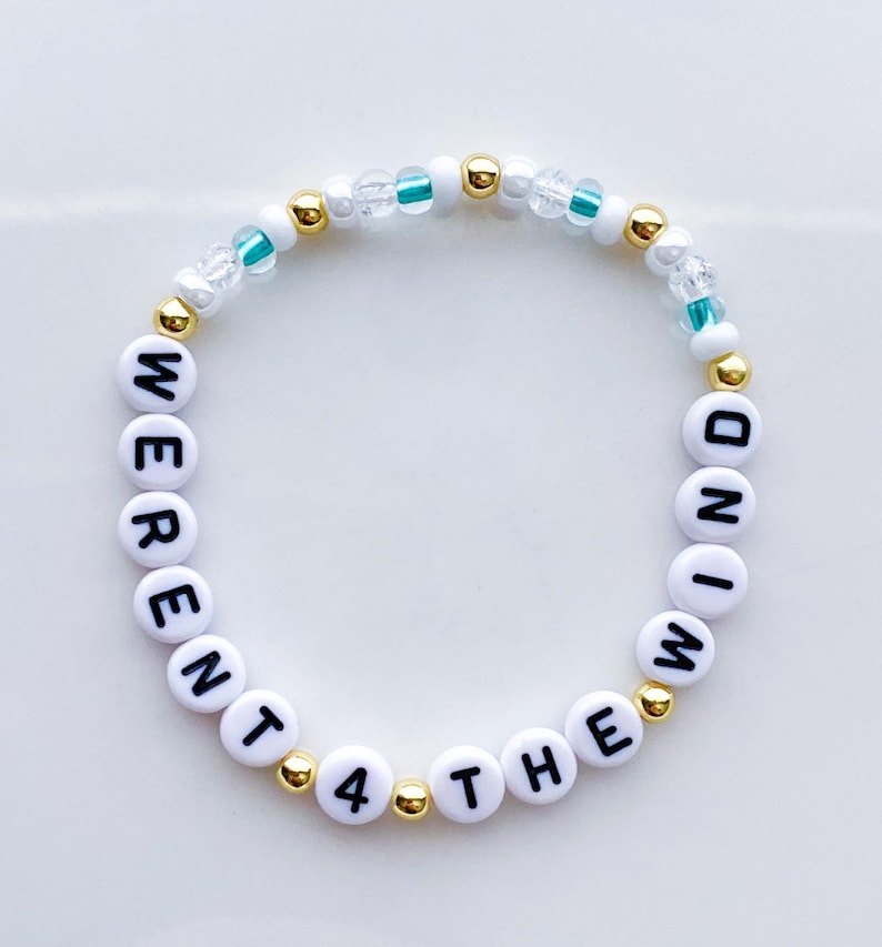 Puede incluir: Una pulsera de cuentas con cuentas de letras blancas que deletrean "WERENT 4 THE WIND". La pulsera tambi&eacute;n presenta cuentas doradas y transparentes con detalles en color turquesa. La pulsera es circular y se encuentra sobre una superficie blanca.