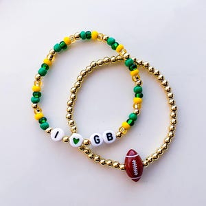 May include: Two gold beaded bracelets. One bracelet has the letters "I", a green heart, and "GB" with a red football charm. The other bracelet has green, yellow, and gold beads.