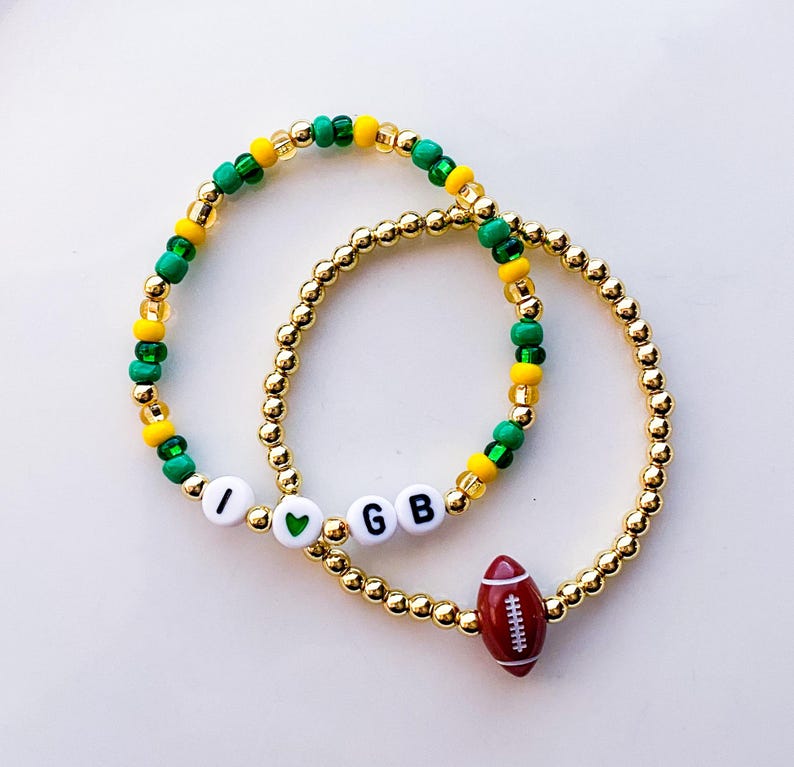 May include: Two gold beaded bracelets. One bracelet has the letters "I", a green heart, and "GB" with a red football charm. The other bracelet has green, yellow, and gold beads.