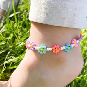 May include: A colorful beaded anklet featuring daisy-shaped beads in shades of pink, yellow, blue, and purple. The anklet is worn on an ankle, with green grass in the background.