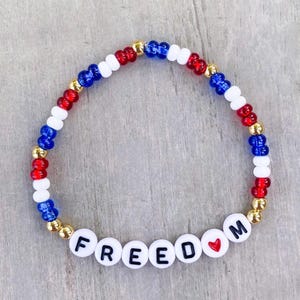 May include: A beaded bracelet with the word "FREEDOM" spelled out in white letter beads. The bracelet features red, white, and blue beads, with gold-colored beads interspersed. A small red heart is between the "D" and "M".