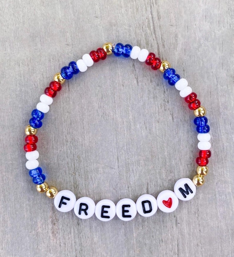 May include: A beaded bracelet with the word "FREEDOM" spelled out in white letter beads. The bracelet features red, white, and blue beads, with gold-colored beads interspersed. A small red heart is between the "D" and "M".