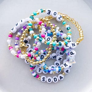 Pode incluir: Uma cole&ccedil;&atilde;o de pulseiras de contas em v&aacute;rias cores, incluindo branco, dourado, rosa e azul-petr&oacute;leo. Algumas pulseiras apresentam contas de letras que soletram nomes e palavras, com pingentes em forma de estrela.