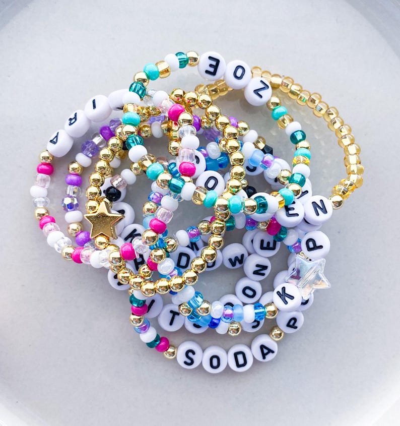 Pode incluir: Uma cole&ccedil;&atilde;o de pulseiras de contas em v&aacute;rias cores, incluindo branco, dourado, rosa e azul-petr&oacute;leo. Algumas pulseiras apresentam contas de letras que soletram nomes e palavras, com pingentes em forma de estrela.