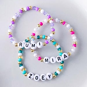 Pode incluir: Tr&ecirc;s pulseiras de contas com contas de letras brancas que soletram "RUMI", "MIRA" e "ZOEY". As pulseiras apresentam uma mistura de contas coloridas em tons de rosa, roxo, dourado, turquesa e branco. As pulseiras est&atilde;o em uma superf&iacute;cie branca.