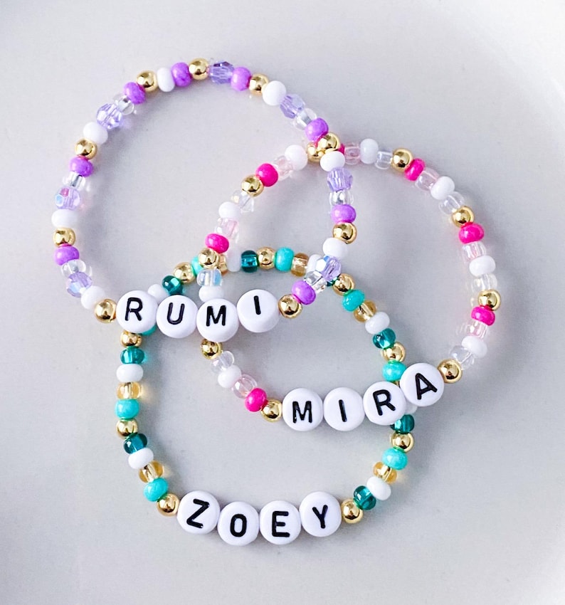 Pode incluir: Tr&ecirc;s pulseiras de contas com contas de letras brancas que soletram "RUMI", "MIRA" e "ZOEY". As pulseiras apresentam uma mistura de contas coloridas em tons de rosa, roxo, dourado, turquesa e branco. As pulseiras est&atilde;o em uma superf&iacute;cie branca.