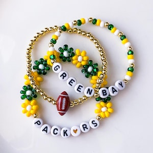 May include: Three beaded bracelets with a Green Bay Packers theme. The bracelets feature gold beads, white letter beads spelling "GREEN BAY PACKERS", and football and flower charms in green, yellow, and white.