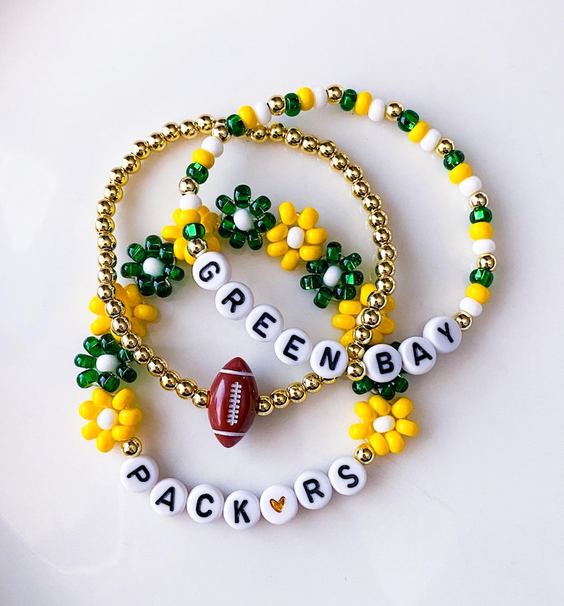 May include: Three beaded bracelets with a Green Bay Packers theme. The bracelets feature gold beads, white letter beads spelling "GREEN BAY PACKERS", and football and flower charms in green, yellow, and white.