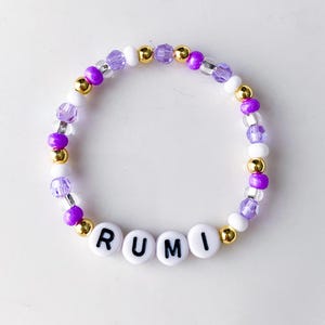 Pode incluir: Uma pulseira de contas com o nome "RUMI" escrito em contas de letras brancas. A pulseira apresenta uma mistura de contas roxas, douradas e brancas. As contas s&atilde;o enfiadas juntas para formar uma forma circular.