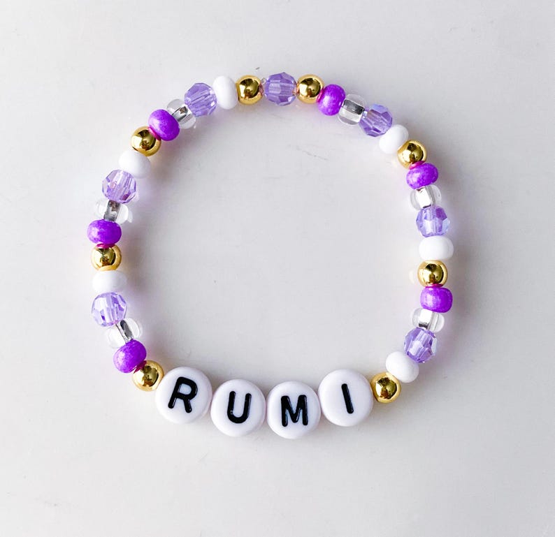 Pode incluir: Uma pulseira de contas com o nome "RUMI" escrito em contas de letras brancas. A pulseira apresenta uma mistura de contas roxas, douradas e brancas. As contas s&atilde;o enfiadas juntas para formar uma forma circular.