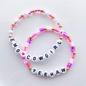 Puede incluir: Dos pulseras de cuentas con las palabras "EMO COWGIRL YEEHAW" escritas con cuentas de letras blancas. Las pulseras presentan cuentas rosas, doradas y transparentes. Una cuenta de coraz&oacute;n rosa est&aacute; entre "EMO" y "COWGIRL".