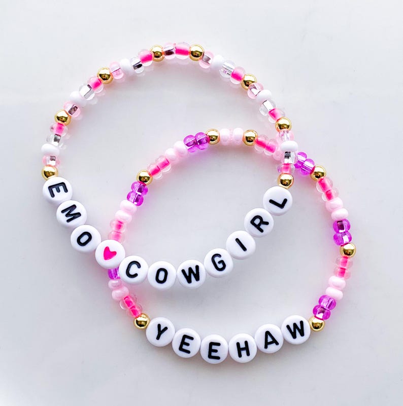 Puede incluir: Dos pulseras de cuentas con las palabras "EMO COWGIRL YEEHAW" escritas con cuentas de letras blancas. Las pulseras presentan cuentas rosas, doradas y transparentes. Una cuenta de coraz&oacute;n rosa est&aacute; entre "EMO" y "COWGIRL".