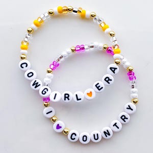 Puede incluir: Dos pulseras de cuentas con las palabras "COWGIRL ERA" y "I LOVE COUNTRY" escritas con cuentas de letras blancas. Las pulseras presentan cuentas doradas, amarillas, rosas y transparentes.