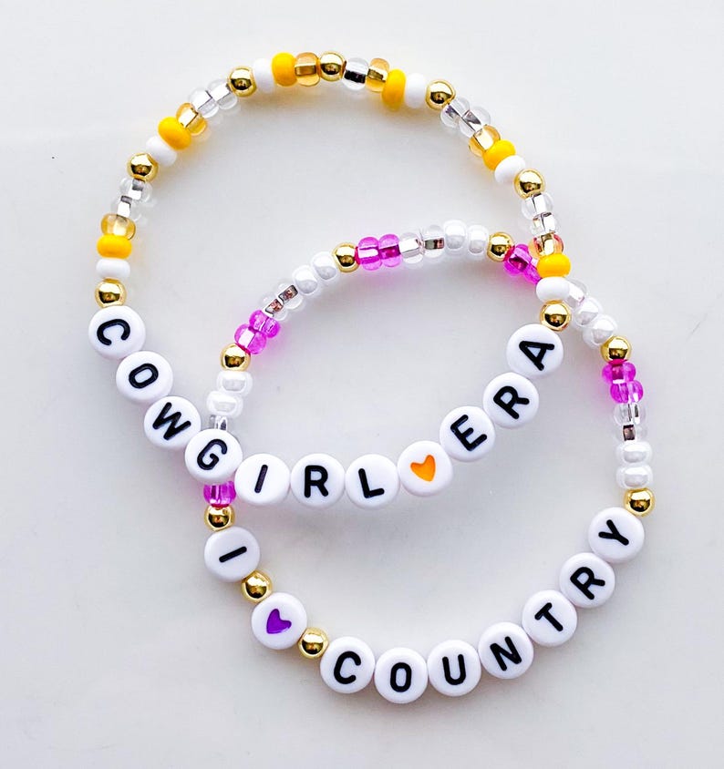 Puede incluir: Dos pulseras de cuentas con las palabras "COWGIRL ERA" y "I LOVE COUNTRY" escritas con cuentas de letras blancas. Las pulseras presentan cuentas doradas, amarillas, rosas y transparentes.