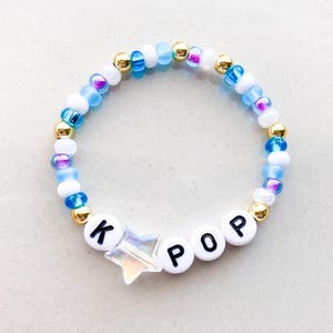 Pode incluir: Uma pulseira de contas com a palavra "POP" escrita em contas brancas. A pulseira apresenta um berloque em forma de estrela e &eacute; feita com uma mistura de contas azuis, brancas, douradas e iridescentes.
