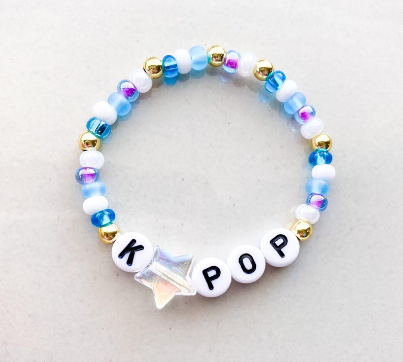 Pode incluir: Uma pulseira de contas com a palavra "POP" escrita em contas brancas. A pulseira apresenta um berloque em forma de estrela e &eacute; feita com uma mistura de contas azuis, brancas, douradas e iridescentes.