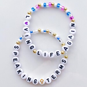 Puede incluir: Dos pulseras de cuentas con letras blancas que deletrean "LIVING LIFE AGAIN" y "SPEAKING TERMS". Las pulseras presentan cuentas doradas y coloridas, incluido un dije de cruz. El fondo es blanco.