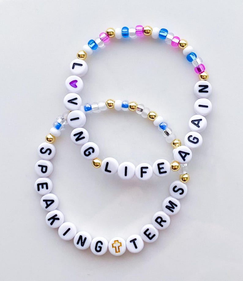 Puede incluir: Dos pulseras de cuentas con letras blancas que deletrean "LIVING LIFE AGAIN" y "SPEAKING TERMS". Las pulseras presentan cuentas doradas y coloridas, incluido un dije de cruz. El fondo es blanco.
