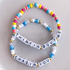 K&ouml;nnte beinhalten: Zwei Armb&auml;nder mit Perlen. Eines tr&auml;gt den Text "THINKIN BOUT ME" und das andere "CHASIN YOU". Die Armb&auml;nder haben bunte Perlen in Rosa, Gelb, Blau und Wei&szlig; mit goldenen Akzenten.