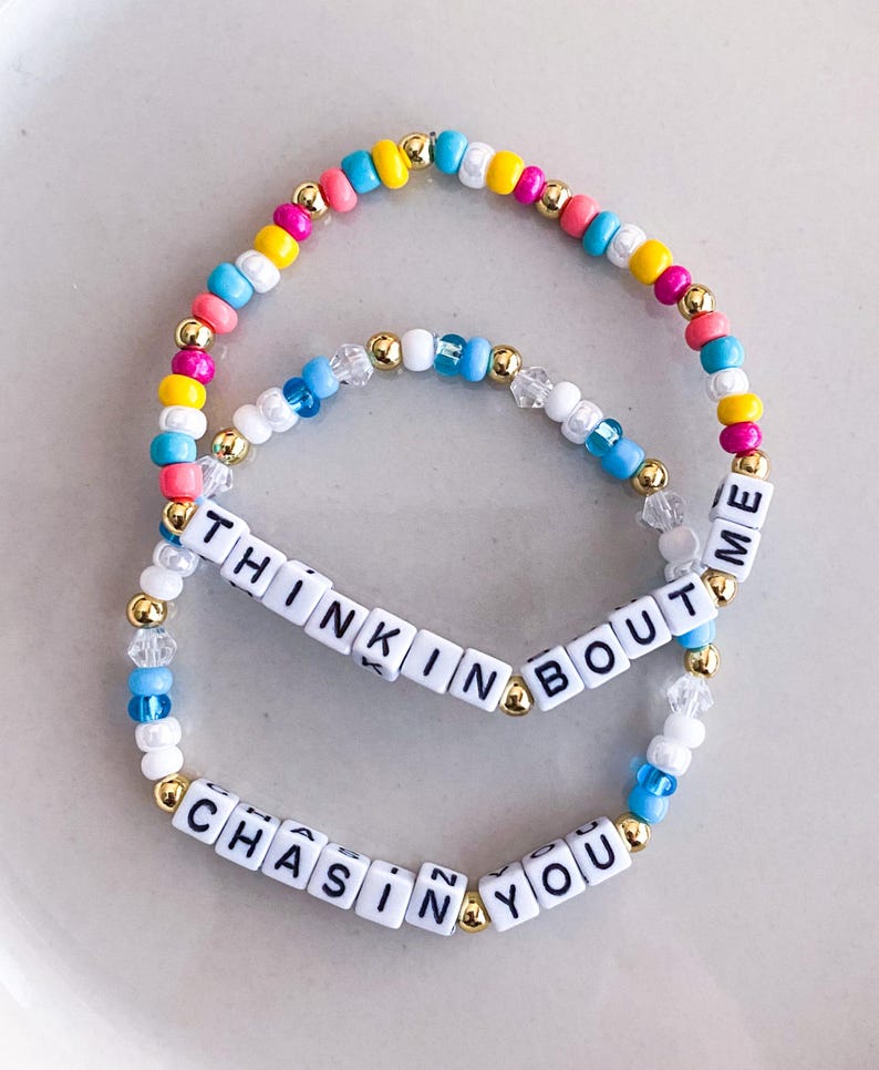 K&ouml;nnte beinhalten: Zwei Armb&auml;nder mit Perlen. Eines tr&auml;gt den Text "THINKIN BOUT ME" und das andere "CHASIN YOU". Die Armb&auml;nder haben bunte Perlen in Rosa, Gelb, Blau und Wei&szlig; mit goldenen Akzenten.