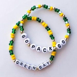 May include: Two beaded bracelets in green, yellow, and gold. One bracelet spells out "GO PACK GO" in white letter beads. The other bracelet spells out "GREEN BAY" in white letter beads. The bracelets are on a white background.