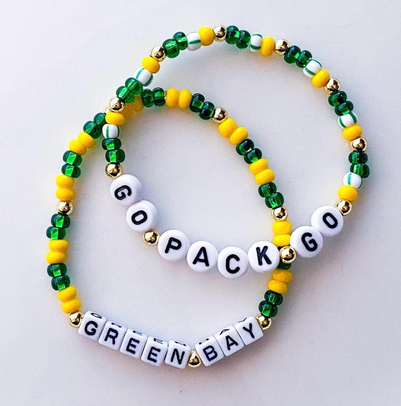 May include: Two beaded bracelets in green, yellow, and gold. One bracelet spells out "GO PACK GO" in white letter beads. The other bracelet spells out "GREEN BAY" in white letter beads. The bracelets are on a white background.