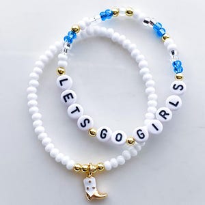 Puede incluir: Dos pulseras de cuentas blancas. Una pulsera deletrea "LETS GO GIRLS" con cuentas de letras negras. La otra pulsera tiene cuentas azules y doradas. Una pulsera tiene un dije dorado de una bota de vaquero blanca.