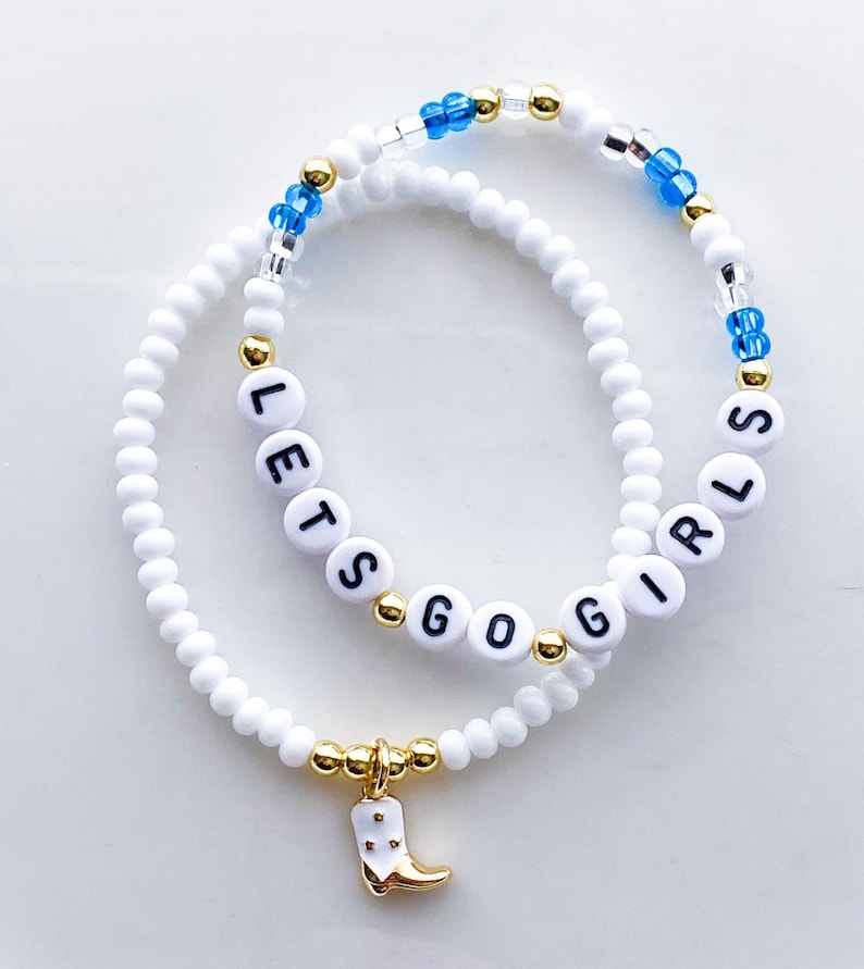 Puede incluir: Dos pulseras de cuentas blancas. Una pulsera deletrea "LETS GO GIRLS" con cuentas de letras negras. La otra pulsera tiene cuentas azules y doradas. Una pulsera tiene un dije dorado de una bota de vaquero blanca.