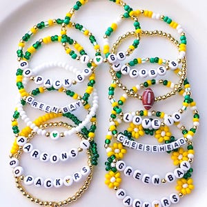May include: A collection of beaded bracelets in green, gold, and white, with text spelling out "GO PACK GO", "GREEN BAY", "JACOBS 8", "LOVE 10", and "CHEESEHEAD". Some bracelets feature football and flower charms.