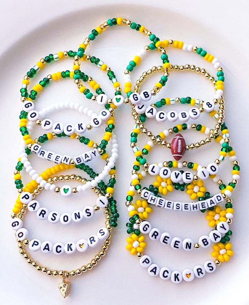 May include: A collection of beaded bracelets in green, gold, and white, with text spelling out "GO PACK GO", "GREEN BAY", "JACOBS 8", "LOVE 10", and "CHEESEHEAD". Some bracelets feature football and flower charms.