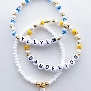 Puede incluir: Tres pulseras de cuentas. Una pulsera deletrea "YLLYLM" con cuentas de letras blancas. Otra pulsera deletrea "DANDELION". Las pulseras tienen cuentas blancas, doradas, amarillas y azules. Un dije dorado de una bota de vaquero blanca cuelga de una pulsera.
