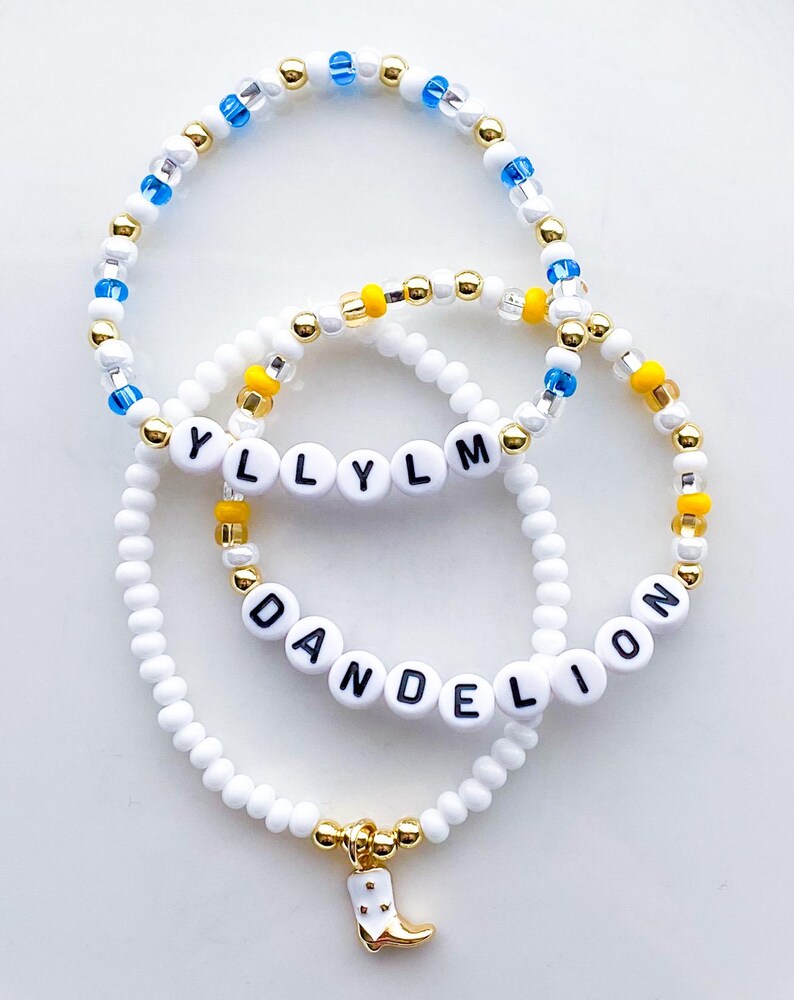 Puede incluir: Tres pulseras de cuentas. Una pulsera deletrea "YLLYLM" con cuentas de letras blancas. Otra pulsera deletrea "DANDELION". Las pulseras tienen cuentas blancas, doradas, amarillas y azules. Un dije dorado de una bota de vaquero blanca cuelga de una pulsera.
