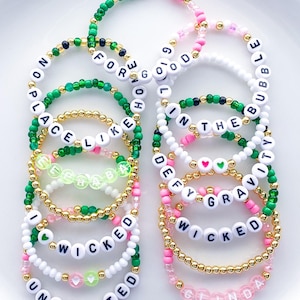 May include: A collection of beaded bracelets in various colors, including green, pink, white, and gold. The bracelets feature letter beads spelling out phrases like "WICKED," "UNLIMITED," and "DEFY GRAVITY." Some bracelets have heart-shaped beads.
