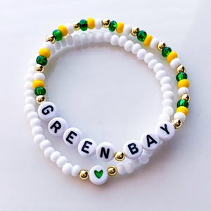 May include: Two white beaded bracelets. One bracelet spells out "GREEN BAY" in black letters on white beads. The other bracelet has green, yellow, and gold beads. A green heart bead is also visible.